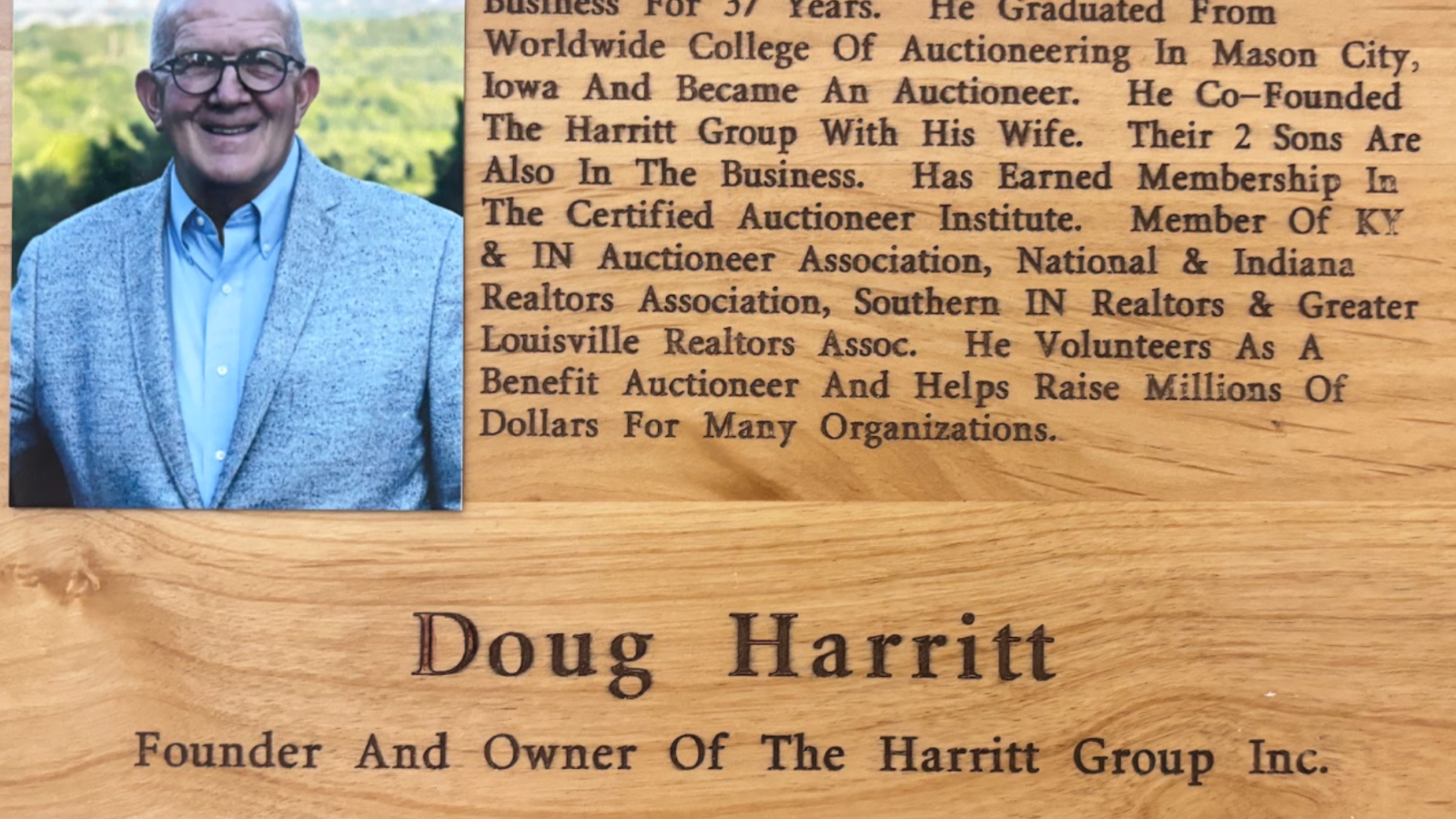 Portrait of a smiling older man wearing glasses and a light blue shirt with a gray blazer, next to a wooden plaque covered in engraved text about his life and achievements.