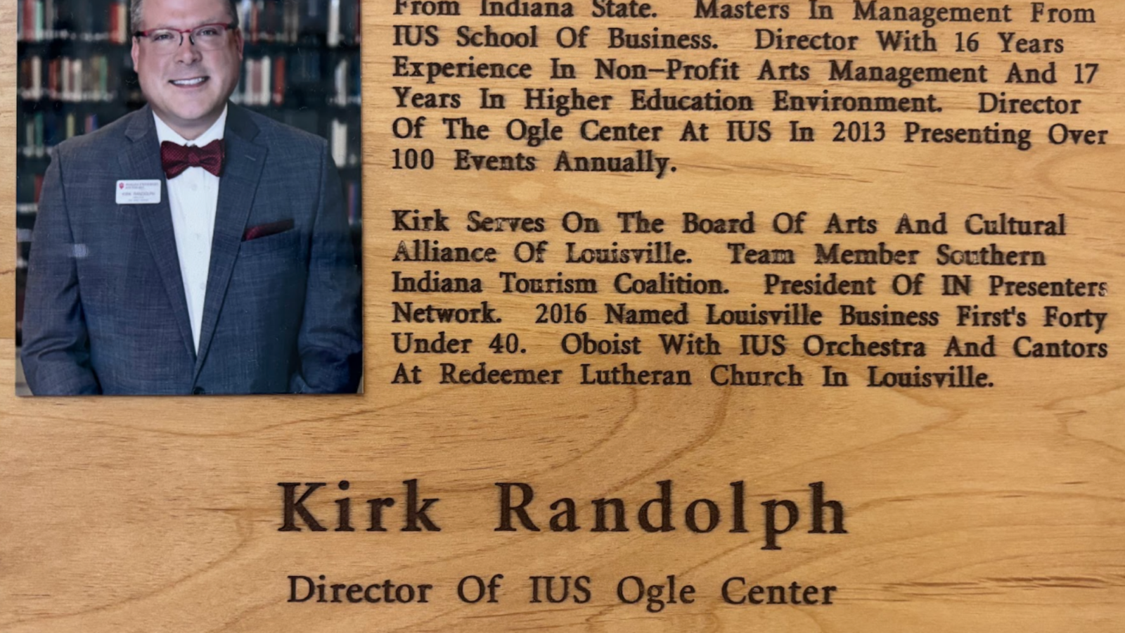 Portrait of a man in a gray suit and red bow tie on a wooden plaque with long biographical text on the right side.
