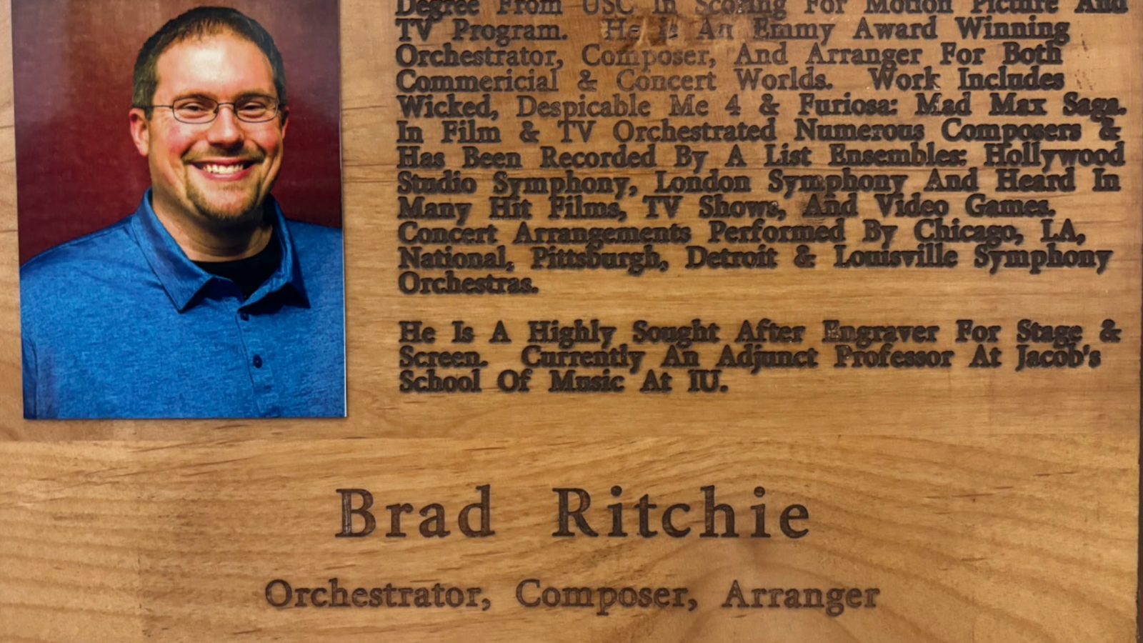 Portrait of a smiling man in a blue shirt on the left, mounted on a wooden plaque with dense biographical text on the right.