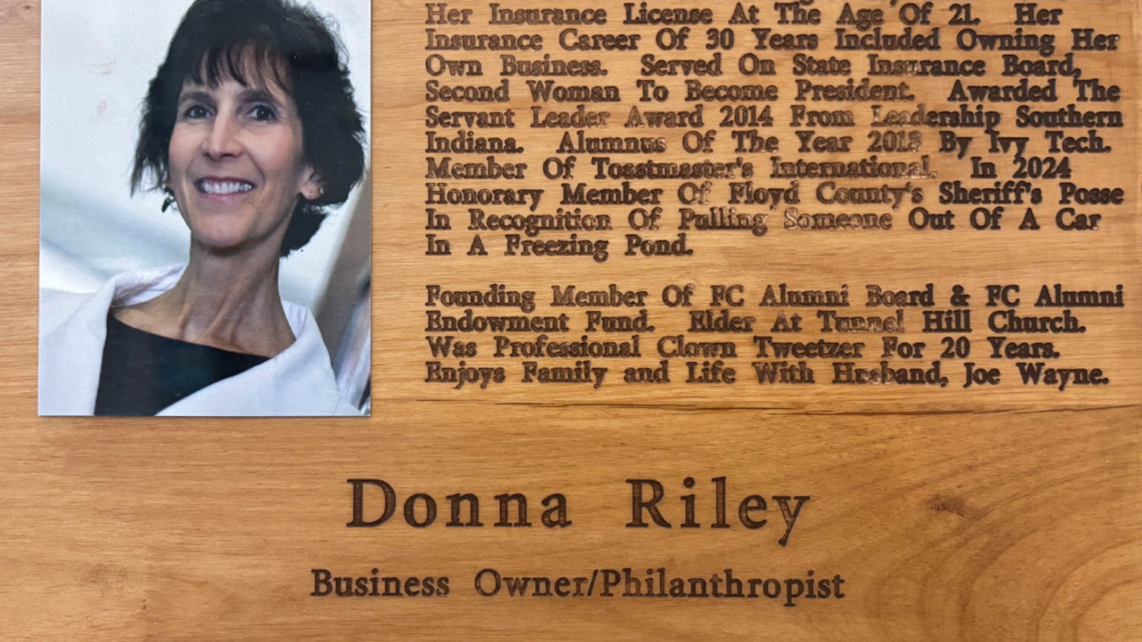 Portrait photo of Donna Riley next to a wooden engraved plaque bearing her name and career achievements, a commemorative display on a wooden background, likely an award plaque.