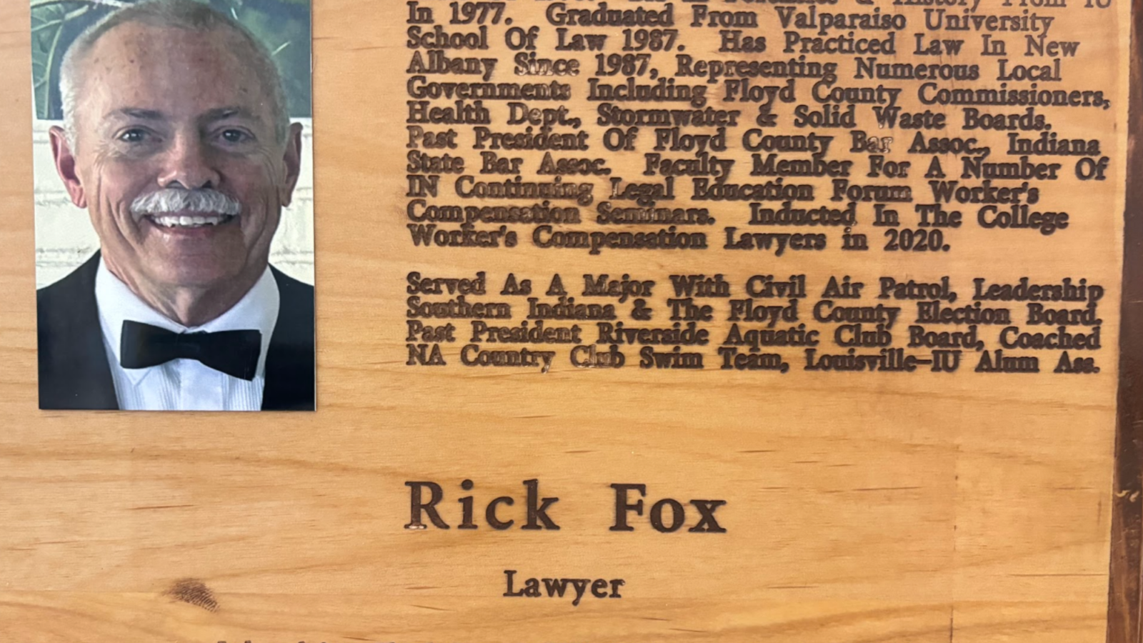Wooden plaque with a left‑side portrait of a man in a tuxedo and a right side filled with dense engraved text about his career achievements.